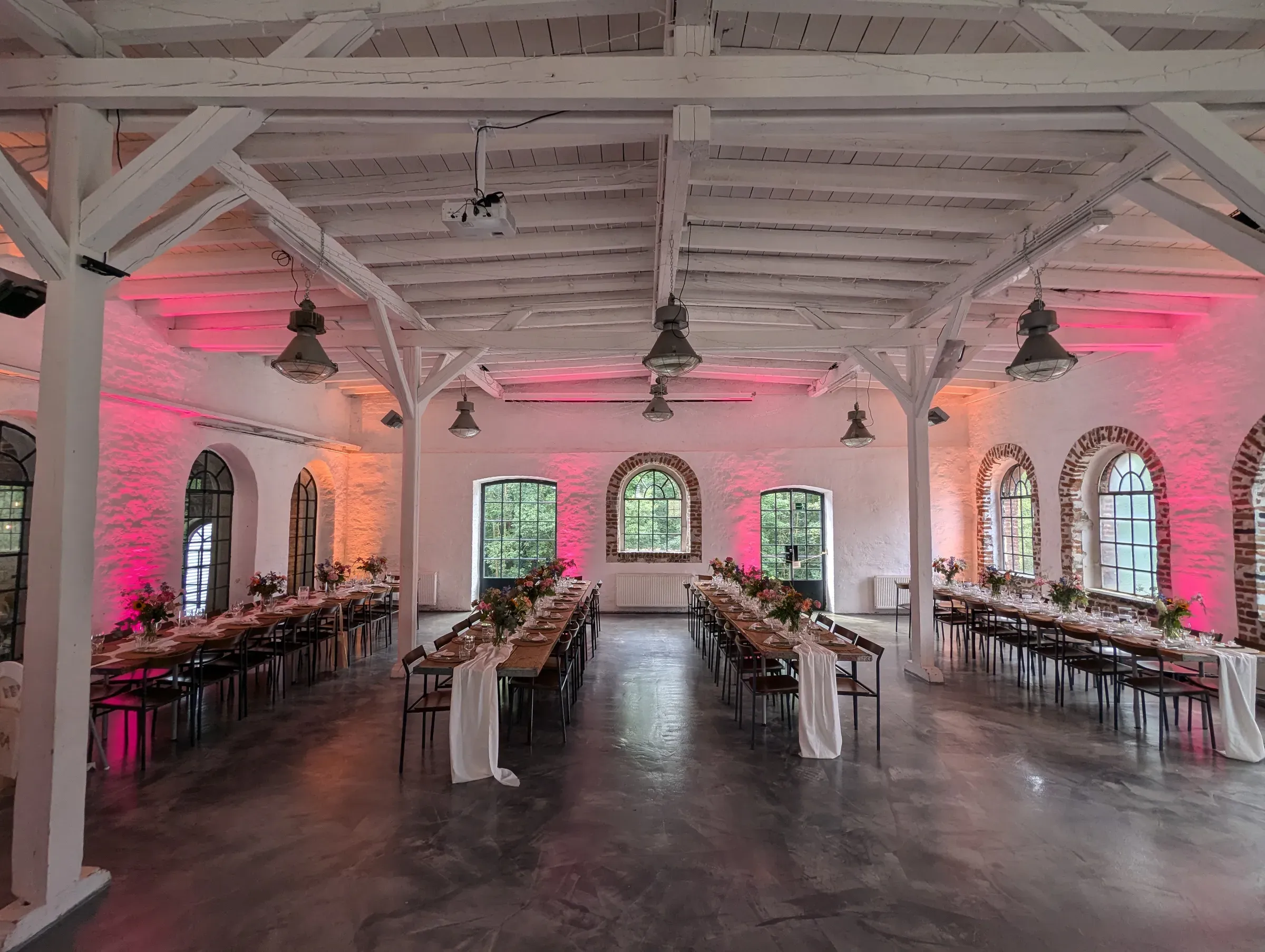 Festsaal mit weißem Holzgebälk und langen gedeckten Tafeln, die Wände leuchten im rosa Licht der Uplights