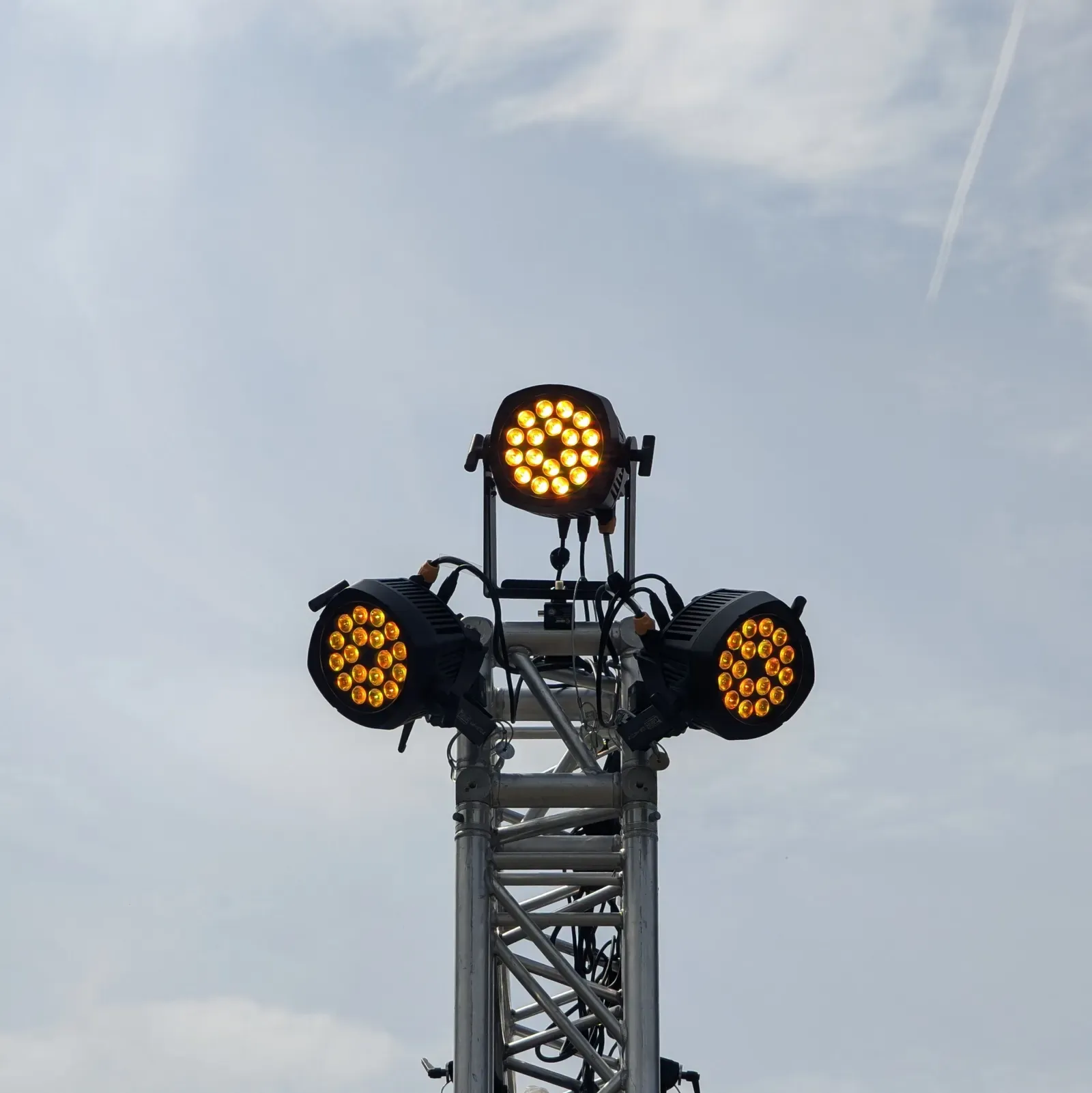 Drei leuchtende LED-Scheinwerfer oben auf einem Traversenturm vor bewölktem Himmel