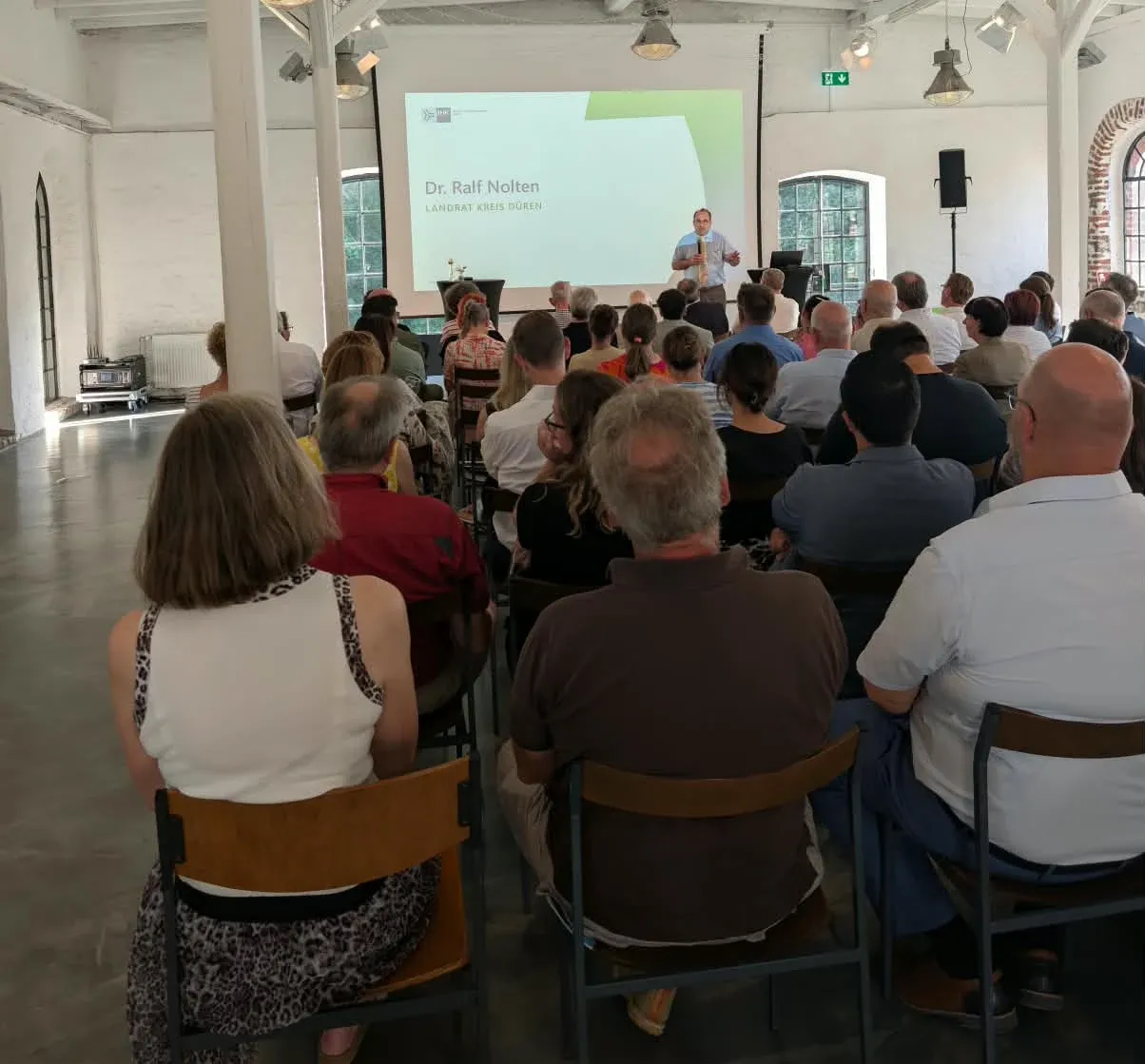 Saal von hinten, Redner mit Mikrofon vor einer großen Leinwand mit Folie